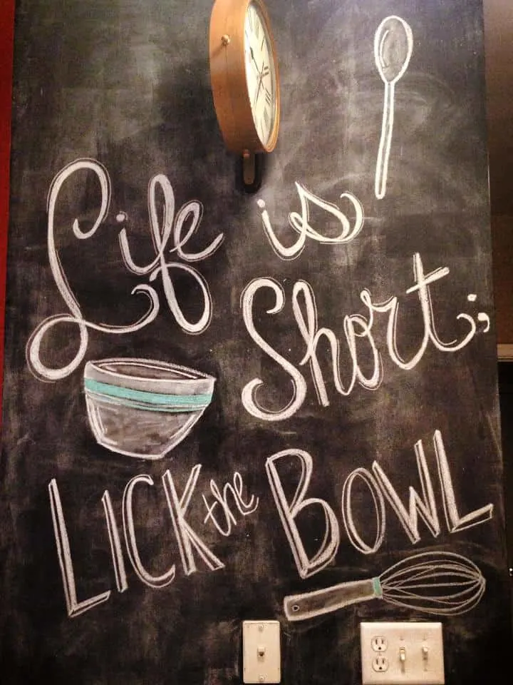 a chalkboard wall with a drawing of a mixing bowl and a text overlay that says, "life is short; lick the bowl"