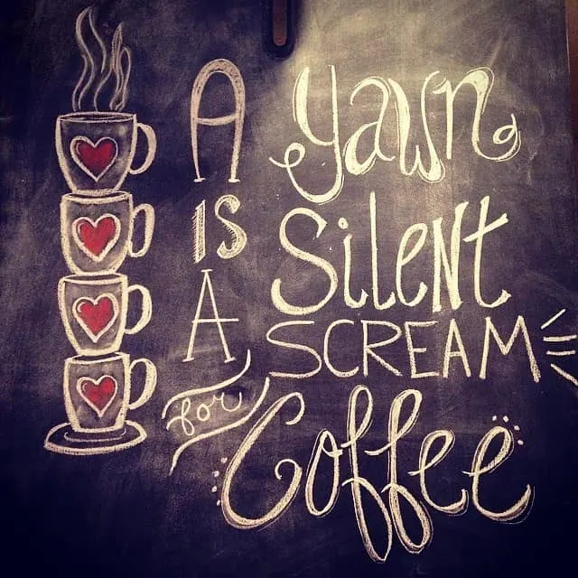 an artistic chalkboard wall with 4 mugs with hearts on them and a text in pretty font that says, "A yawn is a silent scream for coffee". 