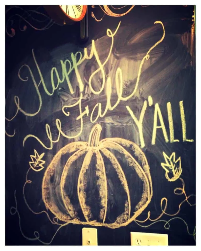 "Happy Fall y'all" written in artistic font on a chalkboard wall in chalk with a chalk drawing of a pumpkin too.