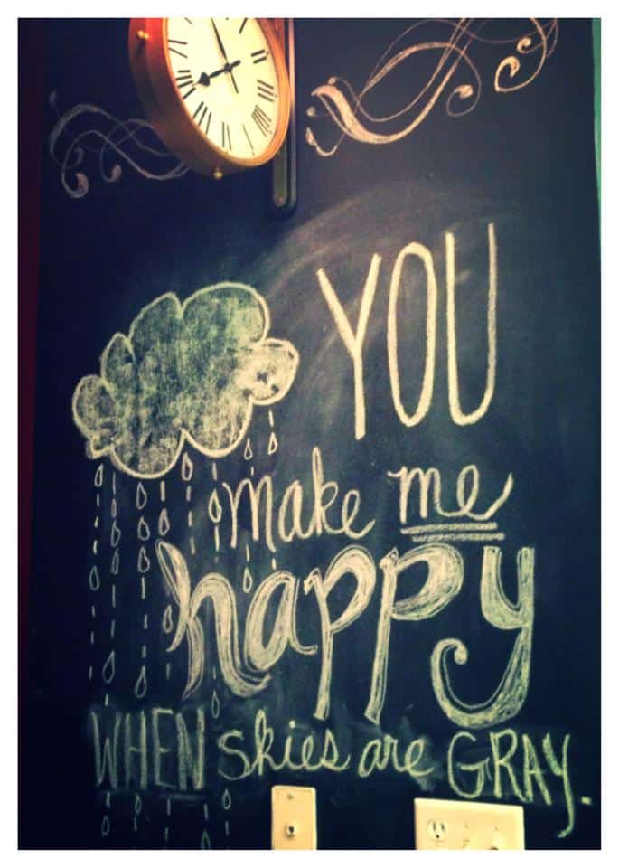 "you make me happy when skies are gray" written in artistic font in chalk on a chalkboard wall. There is also a cloud with rain falling from it, also drawn in chalk.