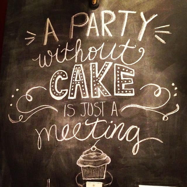 "A party without cake is just a meeting" written in artistic font on a chalkboard wall.