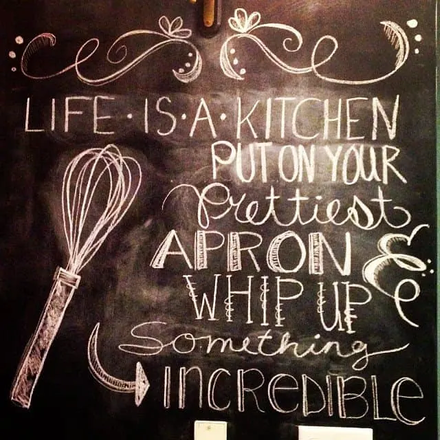 "Life is a kitchen. Put on your prettiest apron and whip up something incredible" written in varrying and unique fonts on a chalkboard wall. There is also a chalk drawing of a whisk. 