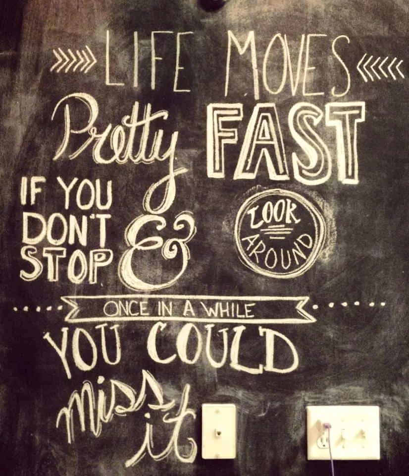 an artistically written quote on a chalkboard wall that says, "life moves pretty fast. If you don't stop and look around once in a while you could miss it".