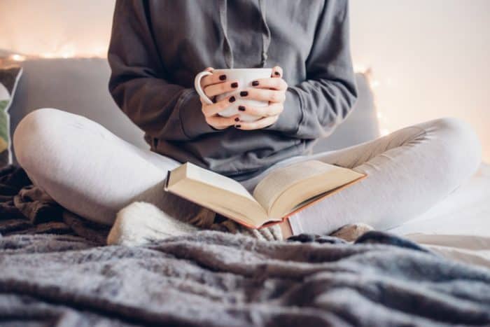 An image of a women sitting cross-legged with a book on her legs and a coffee cup in her hands. The image cuts off at her shoulders.